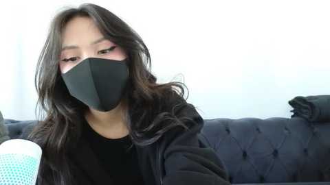 Media: Video of an Asian woman with long, wavy black hair, wearing a black face mask, black hoodie, and sitting on a dark, tufted sofa. She has pink eyeshadow and a neutral expression.