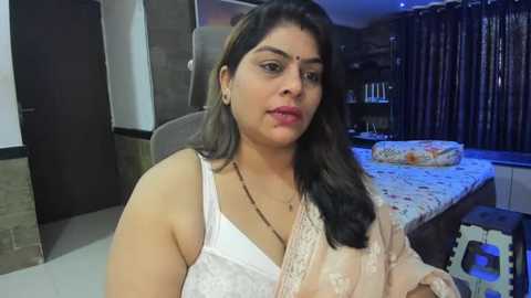 Media: Video of a South Asian woman with medium skin tone, long dark hair, and a bindi, wearing a white lace bra and beige saree. She sits on a brown chair in a dimly lit bedroom with a bed and dark curtains in the background.