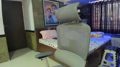 Media: Video of a modern bedroom with a grey mesh office chair facing a wooden bed covered in a floral-patterned sheet, framed family photo on the wall, dark curtains, and brick wall.