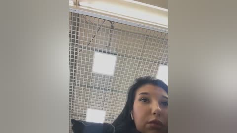 Media: Video of a young Asian woman with long black hair, wearing headphones, sitting in a brightly lit room with a grid-patterned ceiling and fluorescent lights.