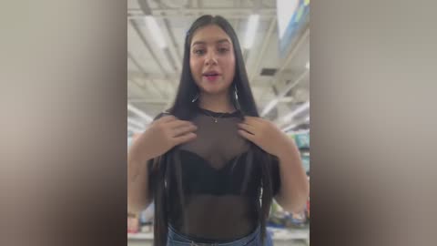 Media: Video of a young Latina woman with long, straight black hair, wearing a sheer black top and denim shorts, standing in a brightly lit, modern grocery store with high ceilings and visible signage.