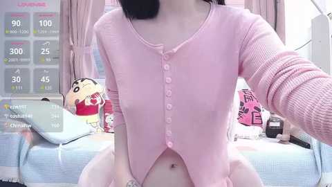 Video of a young woman with pale skin, short black hair, and small breasts, wearing a pink ribbed cardigan, sitting on a bed with pink curtains and a stuffed toy.