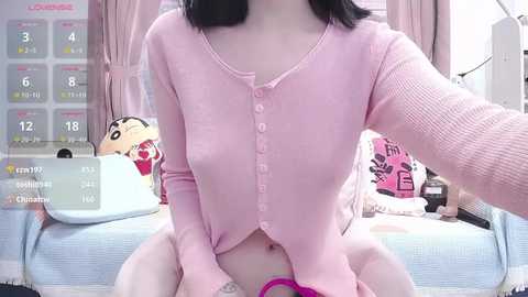 Media: Video of an Asian woman with fair skin, black hair, and medium breasts, wearing a pink cardigan, sitting on a bed with a stuffed animal, calendar, and pink curtains in the background.