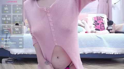 A video of a slim, fair-skinned person wearing a pink, button-up bodysuit, standing in a pastel bedroom. The person's head is cropped out, and the background includes a bed with pink and blue bedding.