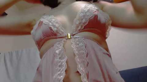 Media: A close-up video of a light-skinned woman with a medium build, wearing a pink, lace-trimmed babydoll lingerie set. The image focuses on her cleavage, nipples, and midriff, with a blurred background of a bedroom setting.