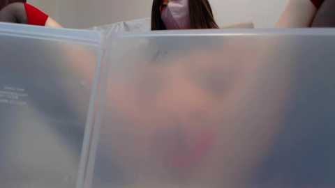 Media: Video of a person with long brown hair and a face mask, partially obscured by a clear plastic folder, in a neutral indoor setting.