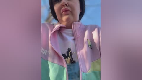 Media: Video of a young girl with short, dark hair, wearing a pastel-colored, multicolored jacket with cartoon characters, outdoors against a bright blue sky.