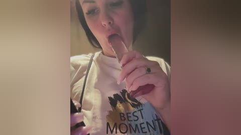 Media: Video of a woman with short dark hair licking an ice cream cone, wearing a white T-shirt with \"BEST MOMENT\" in black letters. The background is blurred and warm-toned.