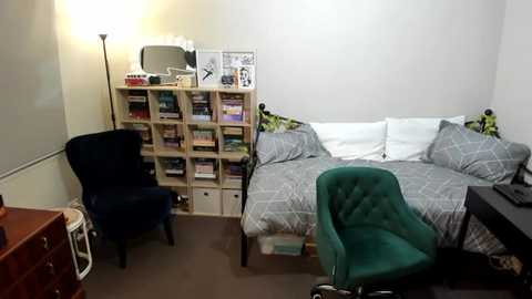 Media: Video of a small, cozy bedroom with a twin bed, green armchair, dark navy armchair, light wood bookshelf, and grey geometric-patterned bedding.