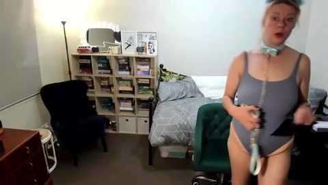 Media: A video of a woman in a gray one-piece swimsuit, holding a phone, standing in a cluttered bedroom with a bed, bookshelf, and dark furniture.