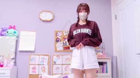 Media: A video of an Asian woman in a cozy, pastel-colored room wearing a face mask, maroon sweatshirt, and white shorts.