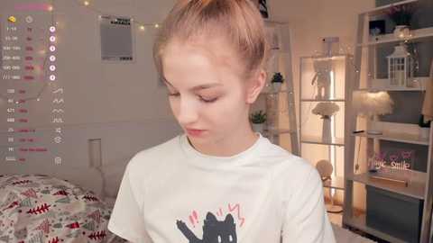 Media: Video of a young, fair-skinned girl with blonde hair in a ponytail, wearing a white T-shirt with a cartoon character, standing in a cozy, warmly lit bedroom with string lights, bookshelves, and a bed in the background.