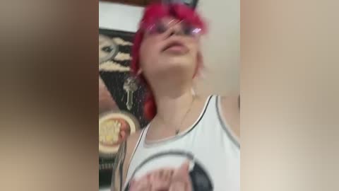 Media: Video of a young woman with pink hair, wearing glasses and a white tank top, holding a pizza. The background shows a wall with a framed poster.