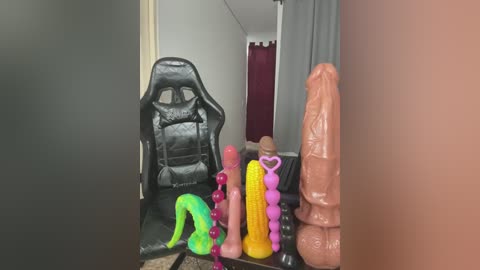 Media: Video of a black gaming chair with a large, realistic penis and various sex toys on a table, including a pink dildo with a purple base, a yellow dildo with a heart-shaped tip, and a purple beaded dildo.