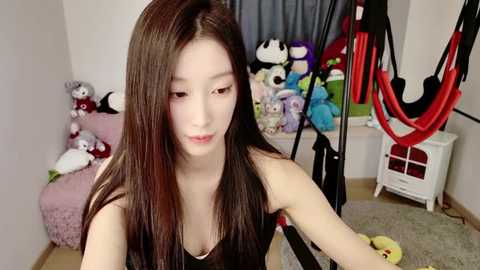 Media: Video of an East Asian woman with long black hair and fair skin, wearing a black top, sitting in a cluttered bedroom filled with stuffed toys and a red and black exercise machine.