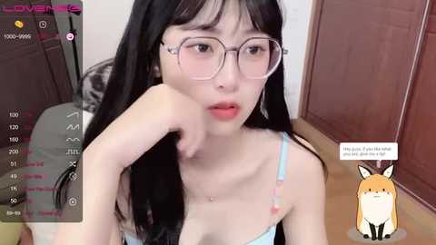 Media: A video of an East Asian woman with long black hair and glasses, wearing a light blue bra, leaning on her elbow, in a cozy room.