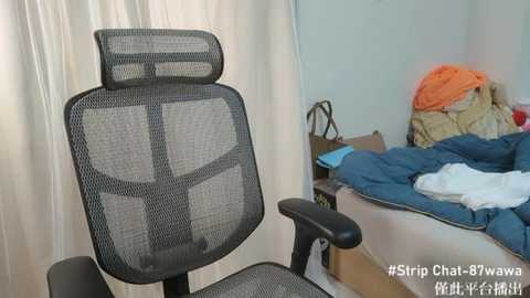 Media: Video of a black mesh office chair with a high backrest in a cluttered bedroom with a messy bed, a pile of clothes, and beige curtains.