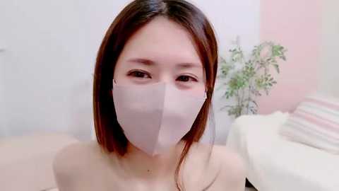 Media: Video of a young Asian woman with long, straight brown hair, wearing a white face mask, in a minimalist room with pale pink and white walls.