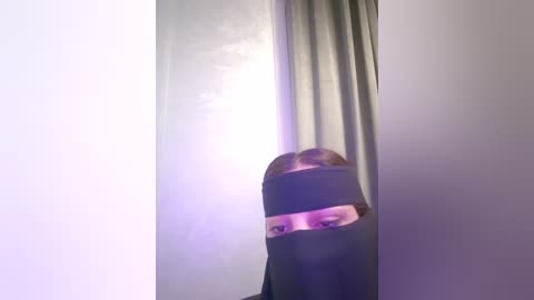 Media: Video of a person with light skin and short, light brown hair, wearing a black niqab covering their face except for their eyes, standing in front of a plain, light-colored curtain.