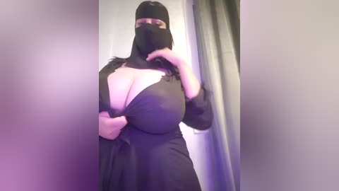 Media: A video of a person in a black burqa with a revealing neckline, exposing large breasts, standing indoors with a blurred background.