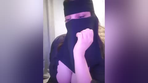 Media: Video of a woman with light skin and long brown hair, wearing a black niqab and a black headscarf, holding a piece of black fabric to her mouth. Background is blurred, with a hint of beige and white colors.