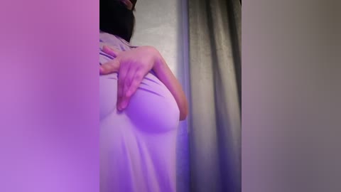 Media: Video of a person with light skin, wearing a white T-shirt, holding their breast, partially covered by a grey curtain. Background is dimly lit with purple and pink hues.