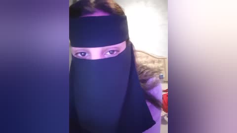 Media: A video of a woman with long brown hair, wearing a black hijab, standing in a bedroom with a bed, beige wall, and a red object in the background.