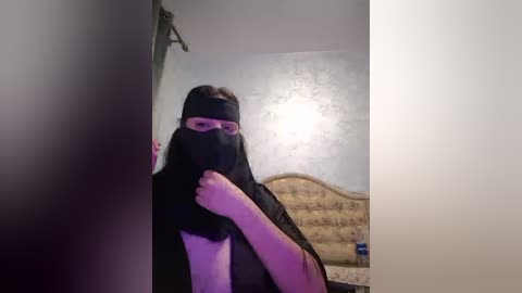 Media: Video of a person in a black mask and glasses, standing in a dimly lit room with a patterned wallpaper and a wooden headboard.