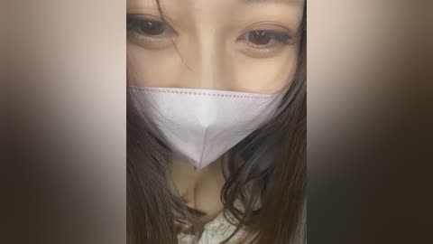 Media: Video of a young woman with long brown hair, wearing a white surgical mask covering her nose and mouth, looking directly into the camera with intense, dark eyes.