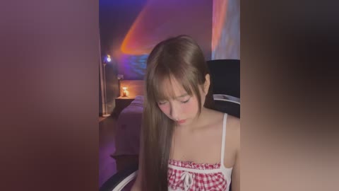 Media: Video of a young Asian girl with long brown hair, wearing a red-and-white checkered dress, sitting on a bed in a dimly lit room with a glowing lamp and colorful wall art.