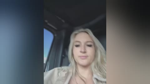 Media: Video of a young Caucasian woman with long blonde hair and fair skin, wearing a beige jacket, seated inside a vehicle with a blue sky visible through the window.