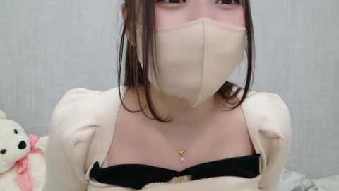 Media: Video of an Asian woman with straight brown hair, wearing a white face mask, black top, and a teddy bear, against a white background.