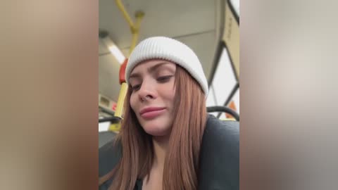 Media: A video of a young Caucasian woman with long, straight brown hair, wearing a white beanie and a black jacket, seated in a bus, eyes closed, looking serene.