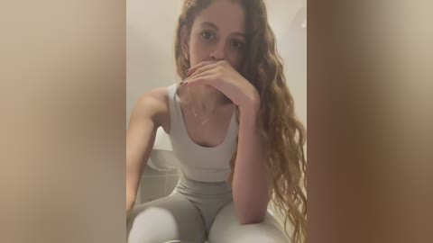 Media: Video of a young woman with curly brown hair, wearing a white sports bra and gray leggings, sitting on a white surface, covering her mouth with her hand.