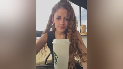 Media: Video of a young, light-skinned woman with long, wavy, brown hair, wearing a black tank top, holding a white Starbucks cup, seated indoors with a blurred background of a caf\u00e9.