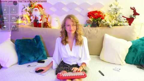 Media: A video of a young blonde woman with wavy hair, wearing a white blouse and black skirt, sitting on a couch with a red keyboard and remote control, surrounded by festive decorations.