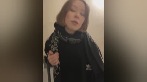 Media: A video shows a young Asian woman with shoulder-length brown hair, wearing a black Adidas jacket and scarf, holding a gun. She has a serious expression. The background is blurred.