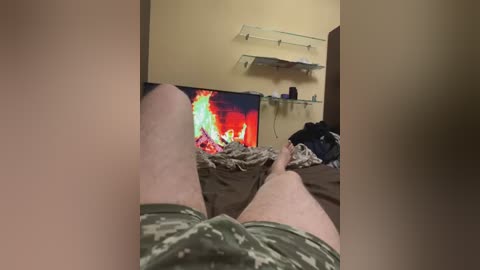 Media: A video of a person's legs in camouflage shorts, lying on a bed with a TV playing a red and yellow flame video. The background features beige walls and a floating shelf with a purple bottle.