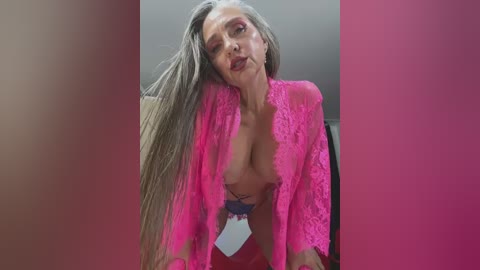 Media: A video of a mature woman with long gray hair, wearing a neon pink lace robe, exposing her breasts and red lingerie, leaning forward provocatively.