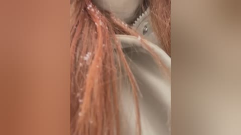 Media: Close-up video of a person's neck with light brown skin, long, reddish-brown hair, and a white jacket zipper partially open. The background is blurred, focusing on the textures and details of the hair and fabric.