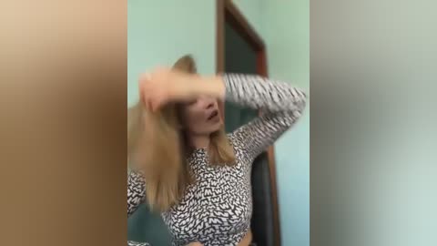 Media: A video of a blonde woman in a leopard-print top, caught mid-motion, covering her face with her hand. She stands in a room with green walls and a wooden door, the image slightly blurred.