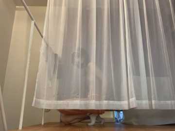 Media: A video showing a child, likely a girl with light skin, crouching under a sheer white curtain in a dimly lit room with wooden floors and beige walls.