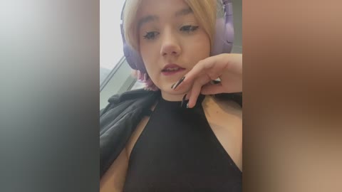 Media: Video of a young, light-skinned woman with blonde hair and large, purple headphones, wearing a black halter top, sitting in a bathroom. She has a neutral expression, with a hand near her mouth.