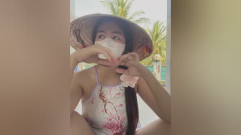 Media: Video of an Asian woman with light brown skin, wearing a floral-patterned dress, a wide-brimmed hat, and a face mask, making a heart shape with her fingers, in front of a palm tree.