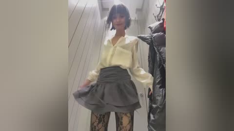 Media: Video of a slender woman with short black hair, wearing a cream blouse, gray skirt, and patterned stockings, standing in a narrow, dimly lit hallway with hanging coats.