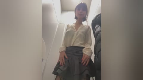 Media: Video of an Asian woman with short black hair and bangs, wearing a white blouse and gray skirt, standing in a narrow, dimly lit hallway.