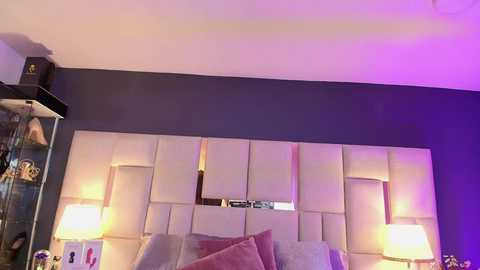 Media: Video of a modern bedroom with a purple-pink lit headboard, beige pillows, and a glass cabinet with decorative items on the left side.