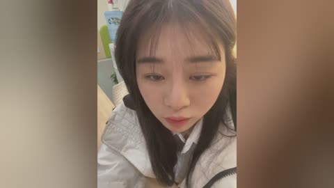 Media: Video of a young East Asian woman with long black hair, wearing a white puffer jacket, looking down with a neutral expression, indoors.