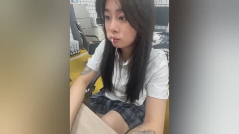 Media: Video of a young Asian woman with long black hair, wearing a white t-shirt and plaid skirt, sitting on a yellow chair in a room with white tiled walls.