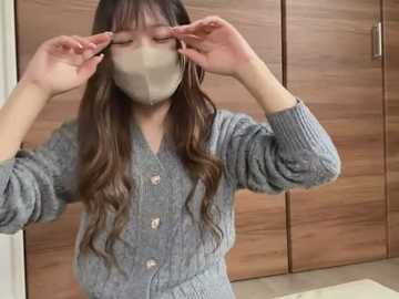 Media: Video of an Asian woman with long, wavy brown hair, wearing a gray cardigan, beige face mask, and large hoop earrings, adjusting her glasses in front of wooden wardrobe doors.